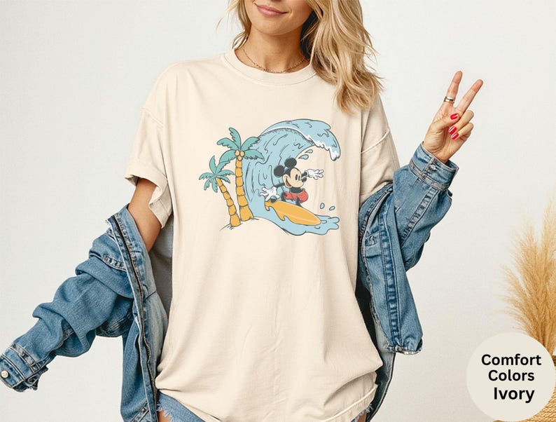 May include: Ivory-colored t-shirt featuring a cartoon Mickey Mouse surfing a wave. The design includes palm trees and a yellow surfboard. The shirt has the words "Comfort Colors Ivory" on the bottom right.