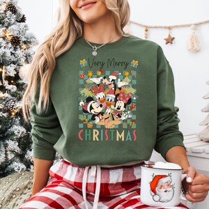 May include: Green sweatshirt with a graphic featuring Disney characters in Santa hats and Christmas trees. The text "Very Merry Christmas" is displayed in a festive font. The person is wearing red and white plaid pants and holding a mug with a Santa Claus design.
