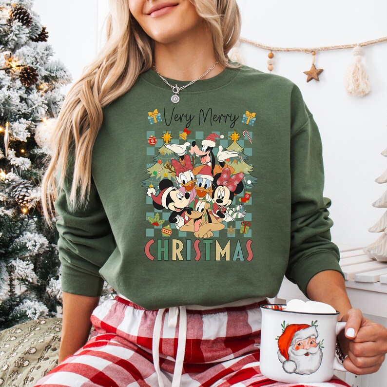 May include: Green sweatshirt with a graphic featuring Disney characters in Santa hats and Christmas trees. The text "Very Merry Christmas" is displayed in a festive font. The person is wearing red and white plaid pants and holding a mug with a Santa Claus design.