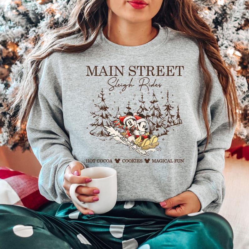 May include: A gray sweatshirt with the text "MAIN STREET Sleigh Rides" and a graphic of Mickey and Minnie Mouse on a sled. The bottom of the shirt reads "HOT COCOA COOKIES MAGICAL FUN".