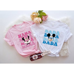 May include: Two t-shirts with a retro design featuring Mickey Mouse and Minnie Mouse. The pink t-shirt says "MAMA" with Minnie Mouse wearing a pink bow. The white t-shirt says "DADA" with Mickey Mouse wearing blue sunglasses.