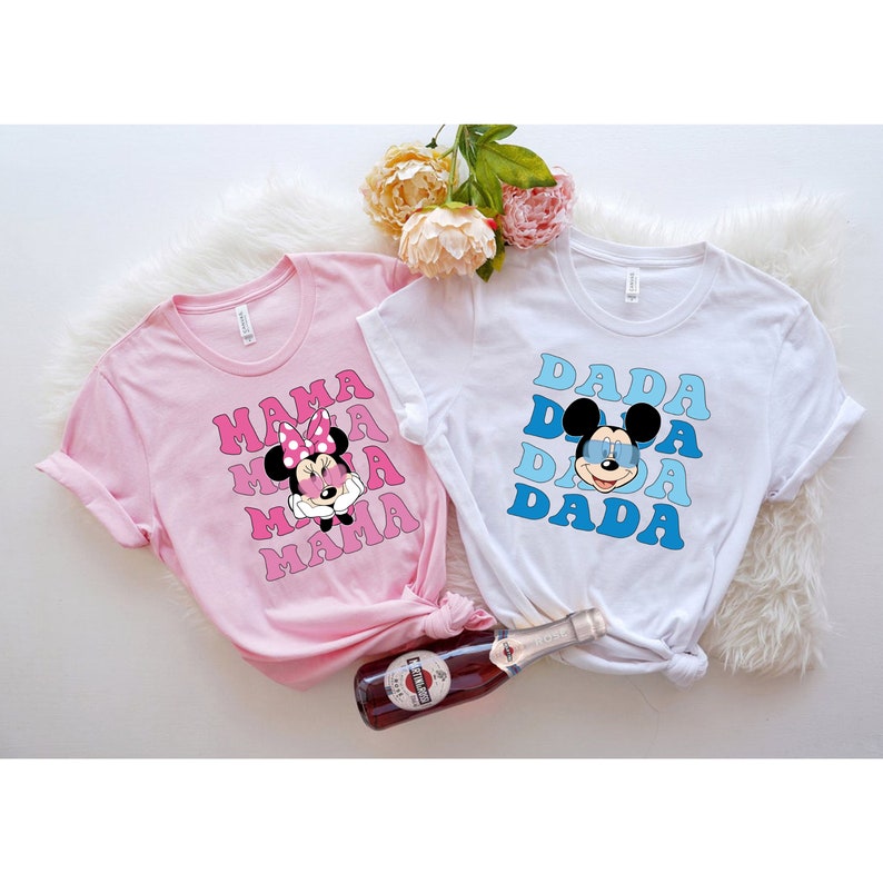 May include: Two t-shirts with a retro design featuring Mickey Mouse and Minnie Mouse. The pink t-shirt says "MAMA" with Minnie Mouse wearing a pink bow. The white t-shirt says "DADA" with Mickey Mouse wearing blue sunglasses.