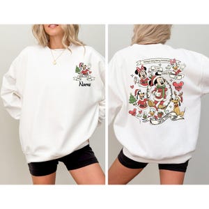 May include: White sweatshirt featuring a festive Disney-themed design. The front displays a small graphic with Mickey Mouse and a Christmas tree, with space for a name. The back showcases a larger illustration of Disney characters celebrating Christmas.