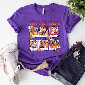 May include: Purple t-shirt with the text "Trick or Treat" in orange and black. The shirt features six cartoon character portraits in Halloween costumes, including Pluto, Mickey Mouse, Donald Duck, Daisy Duck, Minnie Mouse, and Goofy.