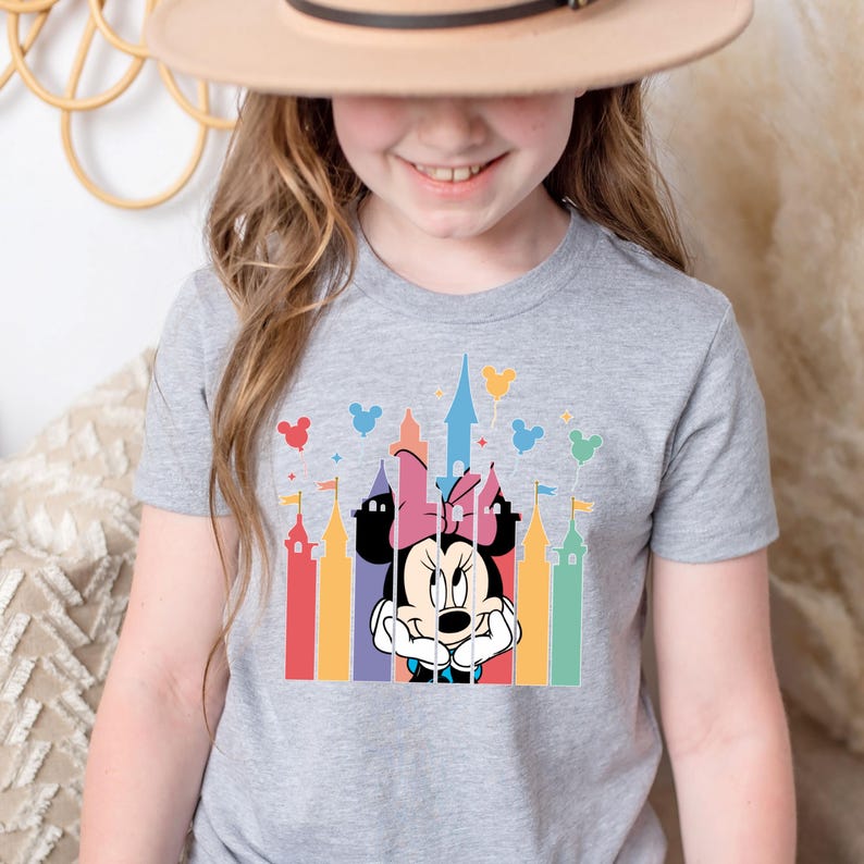 May include: A gray t-shirt with a colorful graphic of Mickey Mouse's girlfriend Minnie Mouse peeking out from behind a silhouette of the iconic Cinderella's Castle at Walt Disney World. Minnie Mouse is wearing a pink bow and has a mischievous expression on her face.