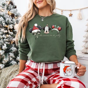 May include: Green sweatshirt with three Mickey Mouse ear designs, each with a Christmas theme. The text "tis the season to be jolly" is printed below the designs. The person is wearing red and white plaid pajama pants and holding a mug with a Santa Claus design.