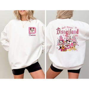 May include: White sweatshirt featuring a pink letter "D" with a cartoon character and the word "Custom" in a script font. The back of the sweatshirt displays a Disneyland graphic with characters and the year 1955 in pink.