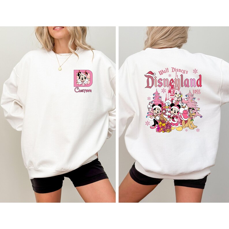 May include: White sweatshirt featuring a pink letter "D" with a cartoon character and the word "Custom" in a script font. The back of the sweatshirt displays a Disneyland graphic with characters and the year 1955 in pink.
