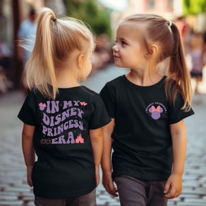 Puede incluir: Dos ni&ntilde;os con camisetas negras con gr&aacute;ficos de tem&aacute;tica de princesas Disney. La parte delantera de una camiseta dice "Disney Princess" con una cabeza de Mickey Mouse morada con un lazo rosa. La parte posterior de la otra camiseta dice "In My Disney Princess Era" con un castillo morado, estrellas rosas y una huella de pata rosa.
