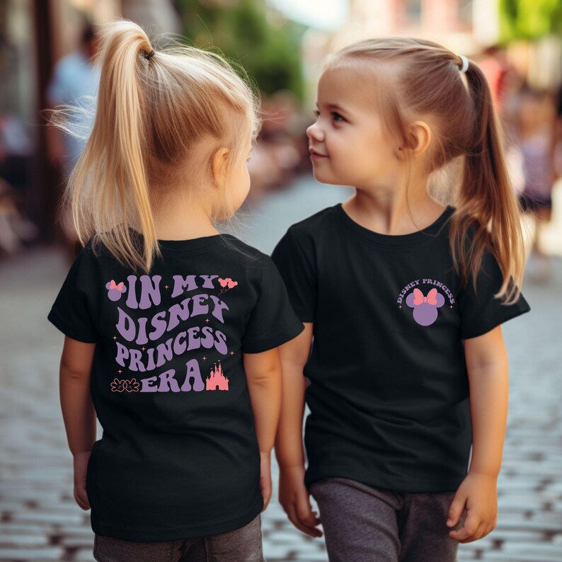 Puede incluir: Dos ni&ntilde;os con camisetas negras con gr&aacute;ficos de tem&aacute;tica de princesas Disney. La parte delantera de una camiseta dice "Disney Princess" con una cabeza de Mickey Mouse morada con un lazo rosa. La parte posterior de la otra camiseta dice "In My Disney Princess Era" con un castillo morado, estrellas rosas y una huella de pata rosa.