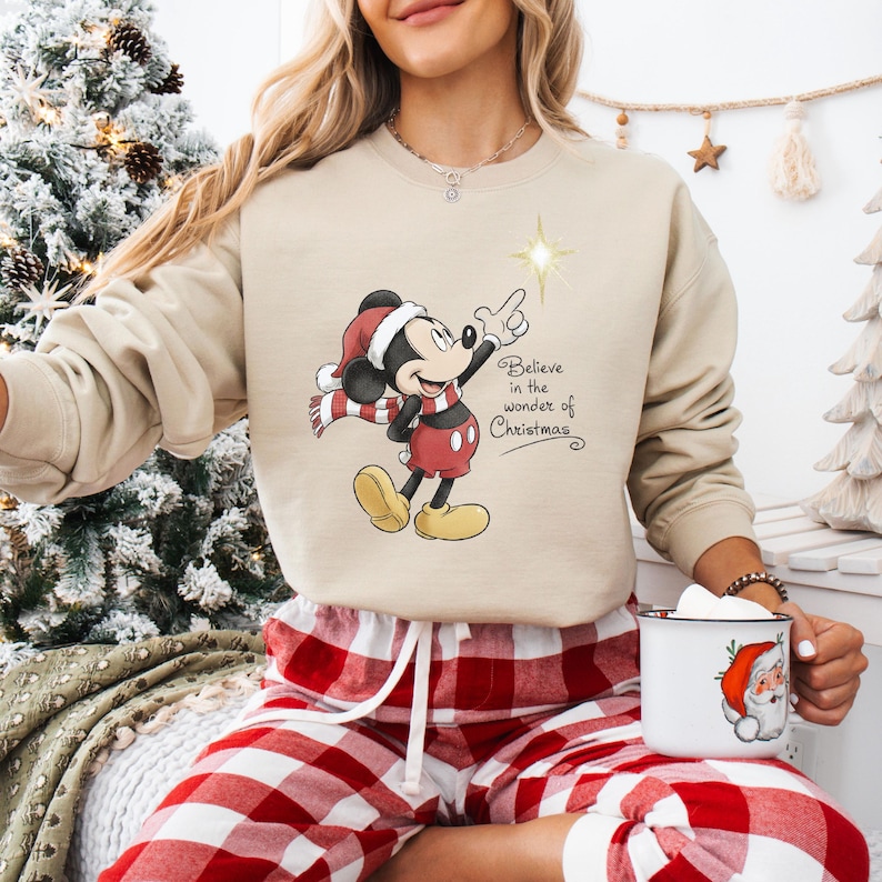 May include: Beige sweatshirt featuring Mickey Mouse in a Santa hat and scarf, pointing at a star. The text "Believe in the wonder of Christmas" is printed on the front. The person is wearing red and white plaid pajama pants.