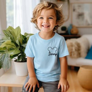 May include: A light blue t-shirt with a black Mickey Mouse head outline and the name "Jerry" in a script font. The shirt is short-sleeved and worn by a smiling child.