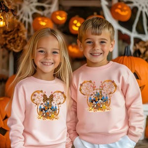 Pode incluir: Duas crian&ccedil;as usando moletons rosa claro combinando. Cada moletom apresenta um design de cabe&ccedil;a do Mickey Mouse com um castelo, personagens e folhagem de outono. O fundo inclui ab&oacute;boras esculpidas e decora&ccedil;&otilde;es de Halloween.