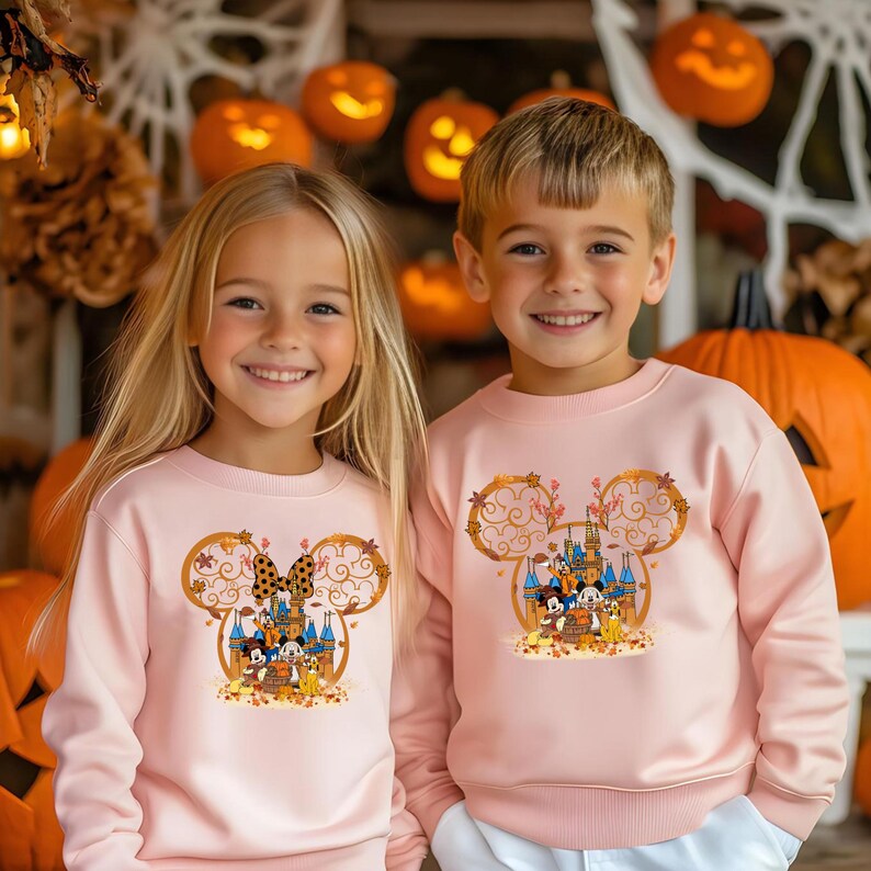 Pode incluir: Duas crian&ccedil;as usando moletons rosa claro combinando. Cada moletom apresenta um design de cabe&ccedil;a do Mickey Mouse com um castelo, personagens e folhagem de outono. O fundo inclui ab&oacute;boras esculpidas e decora&ccedil;&otilde;es de Halloween.