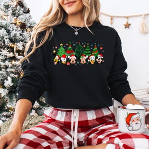 May include: Black sweatshirt featuring a festive design with cartoon characters and Christmas trees. The graphic includes Donald Duck, Mickey Mouse, and other characters wearing Santa hats. The trees are decorated with ornaments and lights. The person is wearing red and white plaid pajama pants and holding a Santa mug.