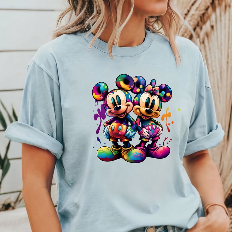 May include: A light blue t-shirt with a colorful graphic of Mickey Mouse and Minnie Mouse holding hands. The graphic is surrounded by colorful paint splatters.