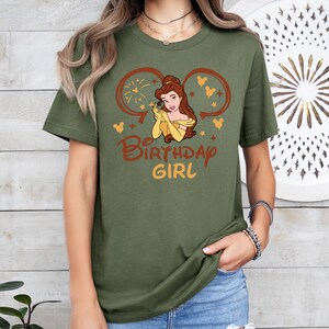 May include: Green t-shirt with a graphic of Belle from Beauty and the Beast wearing a yellow dress and a crown. The graphic is surrounded by Mickey Mouse ears and the text "Be Birthday Girl".