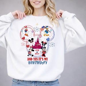May include: White sweatshirt featuring a colorful graphic of Disney characters celebrating the 4th of July. The design includes Mickey and Minnie Mouse, Donald Duck, and Daisy Duck, with fireworks and a castle. Text reads "AND YES IT'S MY BIRTHDAY".