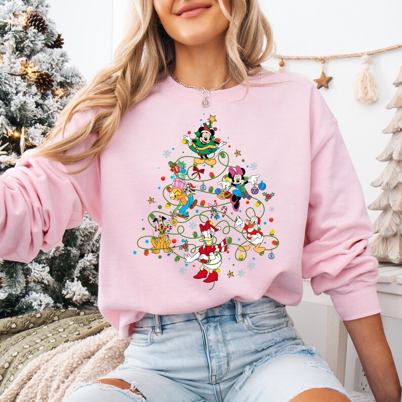 K&ouml;nnte beinhalten: Ein hellrosa Sweatshirt mit einem Weihnachtsbaum-Design aus Disney-Figuren. Micky Maus, Minnie Maus, Donald Duck und Goofy sind abgebildet. Der Baum ist mit Lichtern und Kugeln geschm&uuml;ckt und schafft ein festliches Urlaubs-Thema.