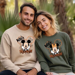 Puede incluir: Una pareja lleva sudaderas a juego de color beige y verde con un dise&ntilde;o de Halloween de Mickey Mouse y Minnie Mouse. Las sudaderas tienen el texto "Boo!" y "Halloween".
