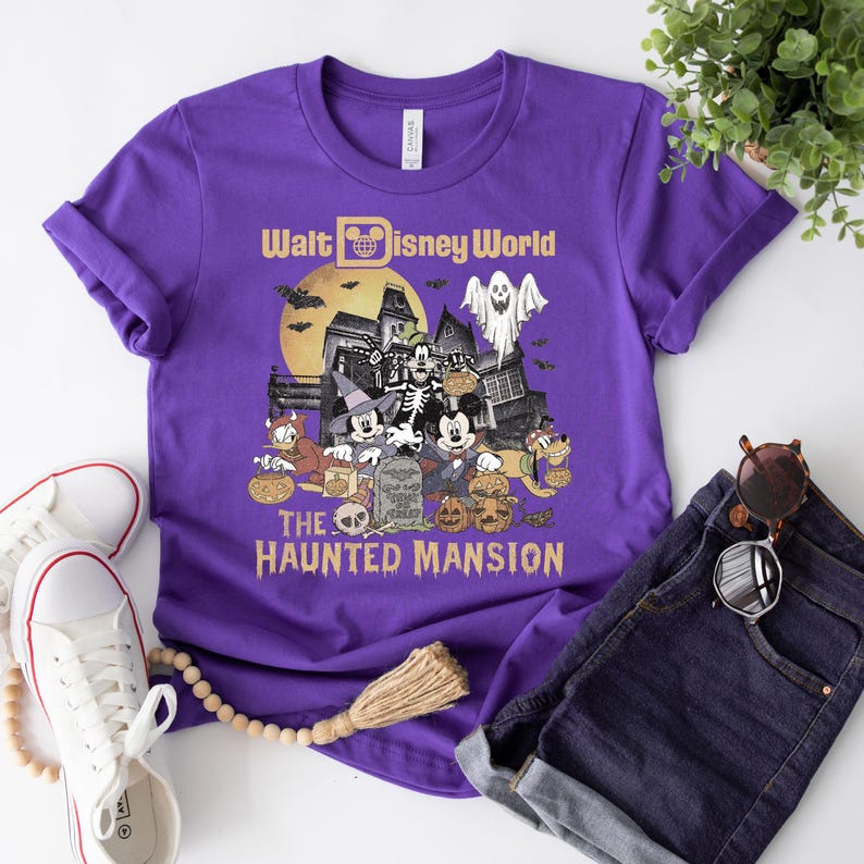 May include: Purple t-shirt with a vintage-style graphic of Mickey Mouse and friends in front of the Haunted Mansion at Walt Disney World. The text "Walt Disney World" and "The Haunted Mansion" are printed on the shirt.