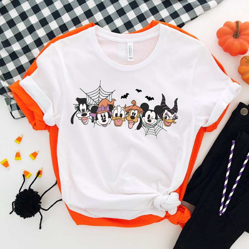 Puede incluir: Camiseta blanca con un gr&aacute;fico de tem&aacute;tica de Halloween con Mickey Mouse, Minnie Mouse, Goofy, Donald Duck, Pluto y Mal&eacute;fica. Los personajes est&aacute;n dispuestos en l&iacute;nea y todos sonr&iacute;en. Hay una telara&ntilde;a y murci&eacute;lagos en el fondo.