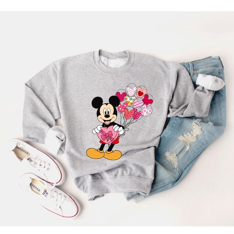 May include: A gray sweatshirt with a graphic of Mickey Mouse holding a heart-shaped box and a bunch of heart-shaped balloons. The balloons are pink, red, and white with a patchwork design.