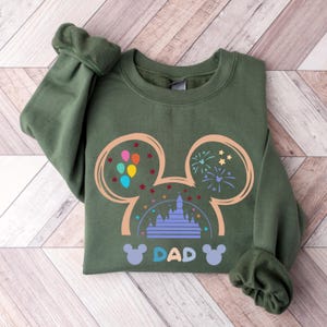 May include: Green sweatshirt with a Mickey Mouse silhouette design. The silhouette includes a castle, balloons, and fireworks. The text "DAD" is printed below the silhouette.