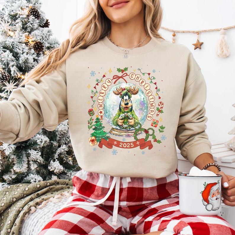 Pode incluir: Camisola bege com um design festivo de globo de neve com uma personagem de desenho animado com chifres. O globo de neve &eacute; rodeado por uma grinalda com as palavras "Merry Christmas" e o ano "2025". A camisola &eacute; combinada com cal&ccedil;as de pijama xadrez vermelhas e brancas.