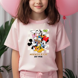 Puede incluir: Camiseta rosa de manga corta con un estampado gr&aacute;fico de Mickey Mouse y sus amigos. El gr&aacute;fico presenta a Mickey Mouse, Minnie Mouse, Donald Duck, Goofy, Pluto y Daisy Duck. El texto "Mickey & Co Est 1928" est&aacute; impreso debajo del gr&aacute;fico.