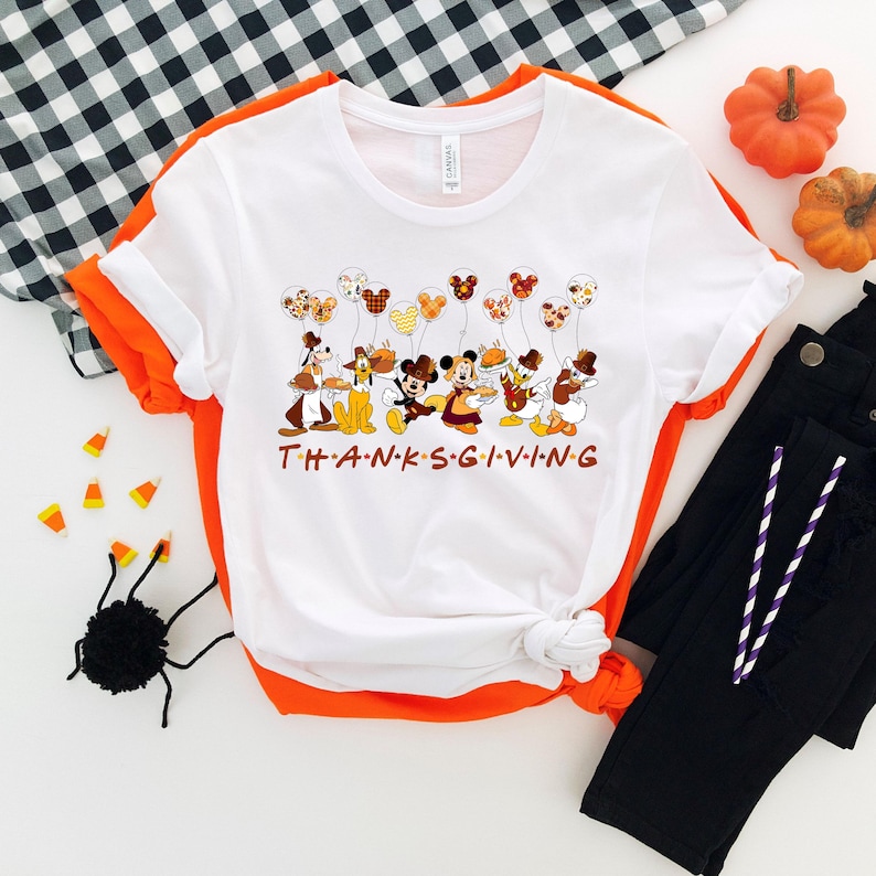 Pu&ograve; includere: T-shirt bianca con grafica a tema Ringraziamento con personaggi dei cartoni animati che tengono cibo e palloncini. La parola "THANKSGIVING" &egrave; stampata in lettere marroni. La maglietta &egrave; abbinata a pantaloni neri e dettagli arancioni.