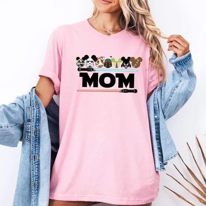 May include: Pink t-shirt featuring a graphic design with Star Wars characters and lightsabers above the word "MOM" in black block letters. The shirt has short sleeves and a relaxed fit.