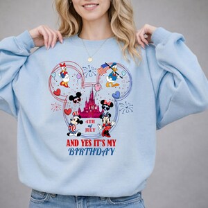 May include: Light blue sweatshirt featuring a Mickey Mouse-shaped design with patriotic characters like Mickey, Minnie, Donald, and Daisy Duck. The design includes fireworks, hearts, and the text "4th of July" and "And Yes It's My Birthday."