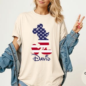 May include: Ivory-colored t-shirt featuring a Mickey Mouse silhouette filled with a patriotic American flag design. The name "David" is printed in navy blue script below the graphic. The shirt is a comfort color.