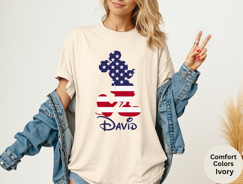 May include: Ivory-colored t-shirt featuring a Mickey Mouse silhouette filled with a patriotic American flag design. The name "David" is printed in navy blue script below the graphic. The shirt is a comfort color.