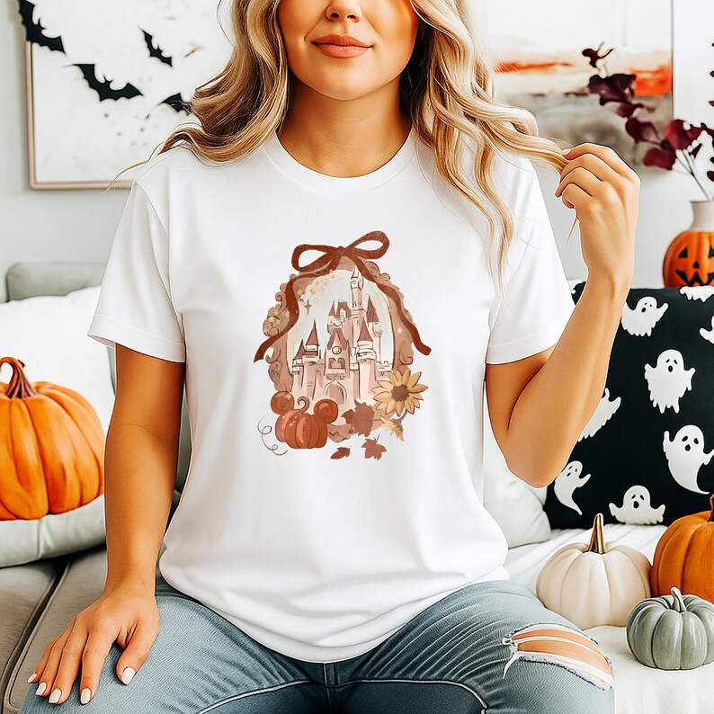 May include: White t-shirt with a fall-themed design. The graphic shows a castle, pumpkins, sunflowers, and leaves, framed by a decorative border. The shirt has a crew neck and short sleeves.