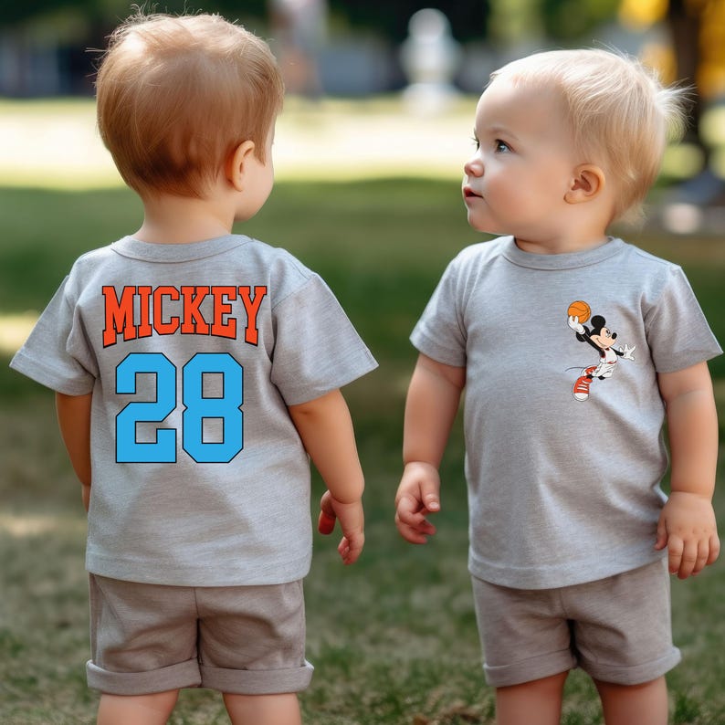 May include: Two toddlers wearing grey t-shirts. One shirt has the text "MICKEY 28" on the back. The other shirt has a cartoon image of Mickey Mouse playing basketball.
