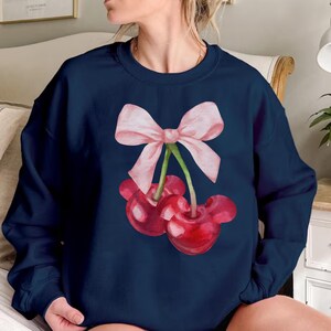 Puede incluir: Una sudadera azul marino con un lazo rosa y dos cerezas rojas. Las cerezas tienen tallos y hojas verdes.