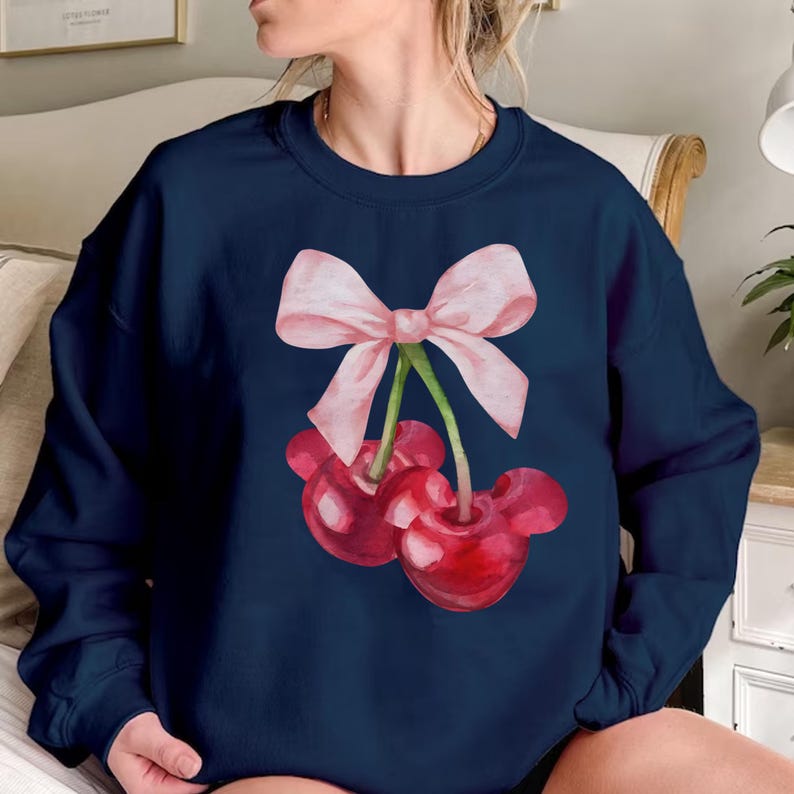 Puede incluir: Una sudadera azul marino con un lazo rosa y dos cerezas rojas. Las cerezas tienen tallos y hojas verdes.