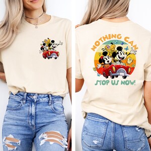 May include: A light beige t-shirt with a vintage-style graphic of Mickey Mouse, Minnie Mouse, and Pluto driving a red car. The graphic is surrounded by a circle with the text "Nothing Can Stop Us Now!" in a retro font.