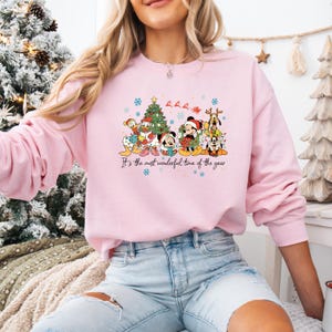 May include: A light pink sweatshirt featuring a festive graphic of Disney characters, including Mickey Mouse, Donald Duck, and Goofy, gathered around a Christmas tree. The text "It's the most wonderful time of the year" is printed below the characters.