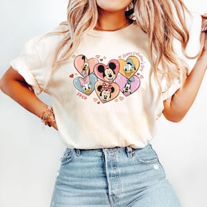 Puede incluir: Una camiseta blanca con un gr&aacute;fico de Mickey Mouse, Minnie Mouse, Donald Duck, Goofy y Pluto en corazones. El texto "Happy Valentine's Day" y "XOXO" tambi&eacute;n est&aacute;n en la camiseta.