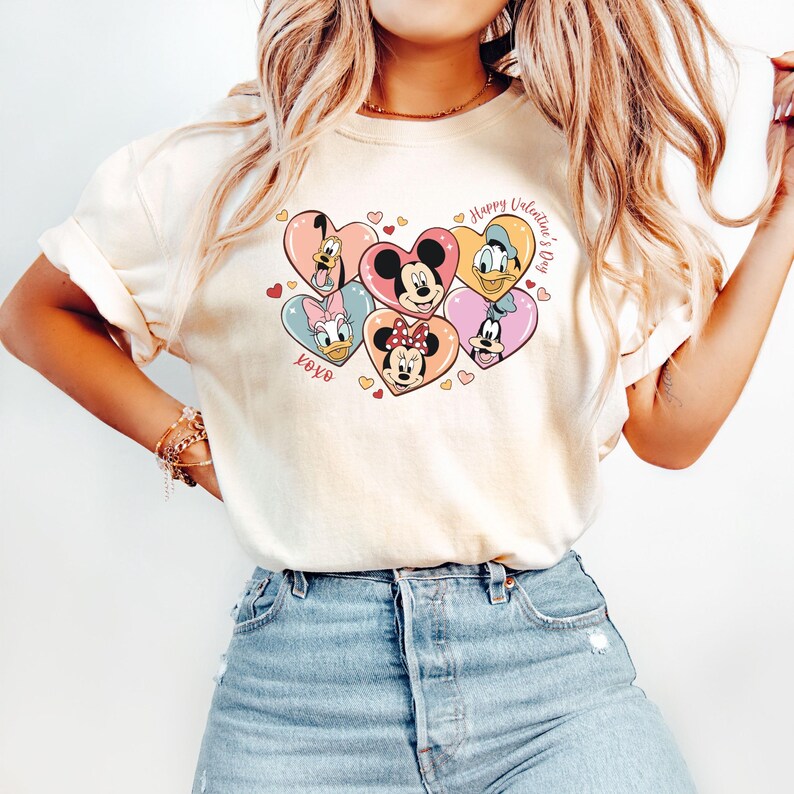 Puede incluir: Una camiseta blanca con un gr&aacute;fico de Mickey Mouse, Minnie Mouse, Donald Duck, Goofy y Pluto en corazones. El texto "Happy Valentine's Day" y "XOXO" tambi&eacute;n est&aacute;n en la camiseta.