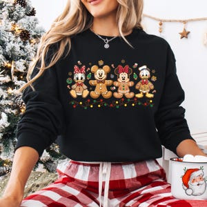 May include: Black sweatshirt featuring gingerbread versions of Disney characters: Daisy Duck, Mickey Mouse, Minnie Mouse, and Donald Duck. The gingerbread figures are decorated with icing and candy, and are arranged above a string of Christmas lights.