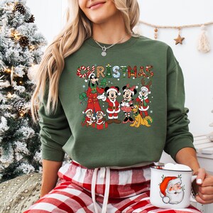 May include: Green Christmas sweatshirt featuring a festive graphic of Disney characters, including Mickey and Minnie Mouse, and the word "CHRISTMAS" in a plaid design. The sweatshirt is paired with red and white plaid pajama pants.