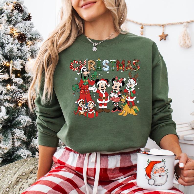 May include: Green Christmas sweatshirt featuring a festive graphic of Disney characters, including Mickey and Minnie Mouse, and the word "CHRISTMAS" in a plaid design. The sweatshirt is paired with red and white plaid pajama pants.