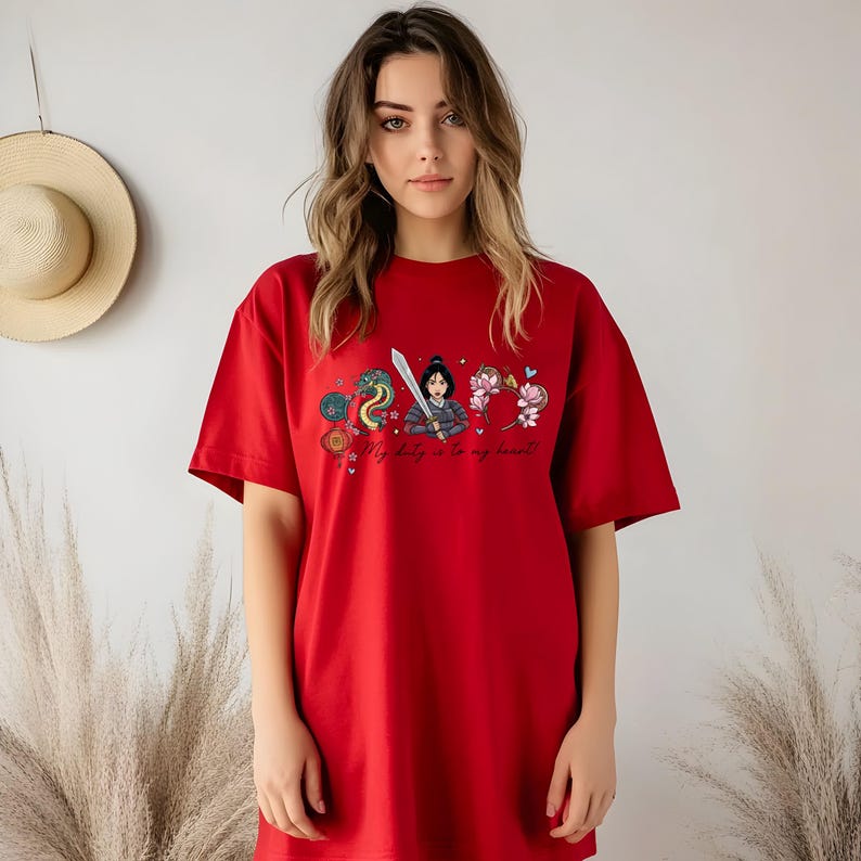 May include: A red t-shirt with a graphic design featuring a warrior woman, a dragon, flowers, and the text "My duty is to my heart!". The design incorporates blue, pink, and gold. The shirt has a relaxed, oversized fit.