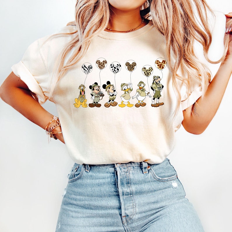 Pode incluir: Uma camiseta branca com um gr&aacute;fico de Mickey Mouse, Donald Duck, Goofy e Pluto vestidos com roupas de saf&aacute;ri e segurando bal&otilde;es com estampa de animais.