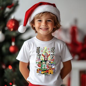 May include: A child wearing a white t-shirt with a colorful Christmas graphic featuring cartoon characters. The child is also wearing a red Santa hat with a white pom-pom. A Christmas tree with red ornaments is in the background.