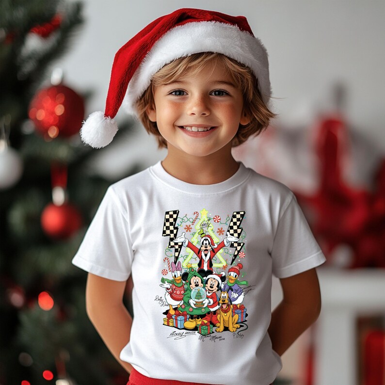 May include: A child wearing a white t-shirt with a colorful Christmas graphic featuring cartoon characters. The child is also wearing a red Santa hat with a white pom-pom. A Christmas tree with red ornaments is in the background.